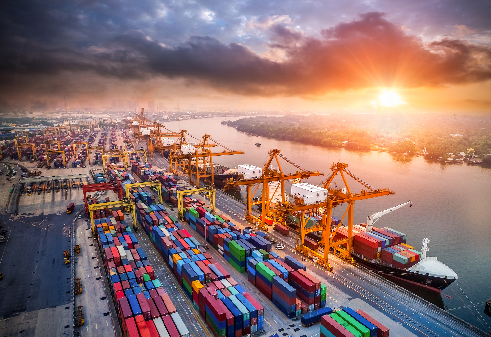 Sunrise over bustling port with cargo containers and cranes by the river.