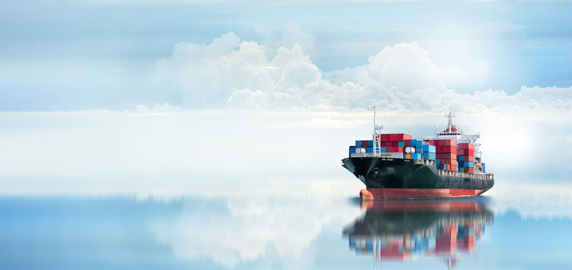 Cargo ship with colorful containers reflected on calm ocean water, under cloudy sky.